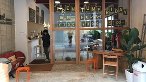 Interior de una cafetería con techo de madera, una puerta de cristal con letreros y un área de estar con mesas y sillas.