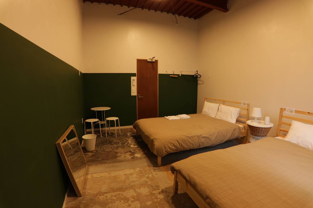 Une petite pièce faiblement éclairée, avec deux lits, une petite table et une chaise.