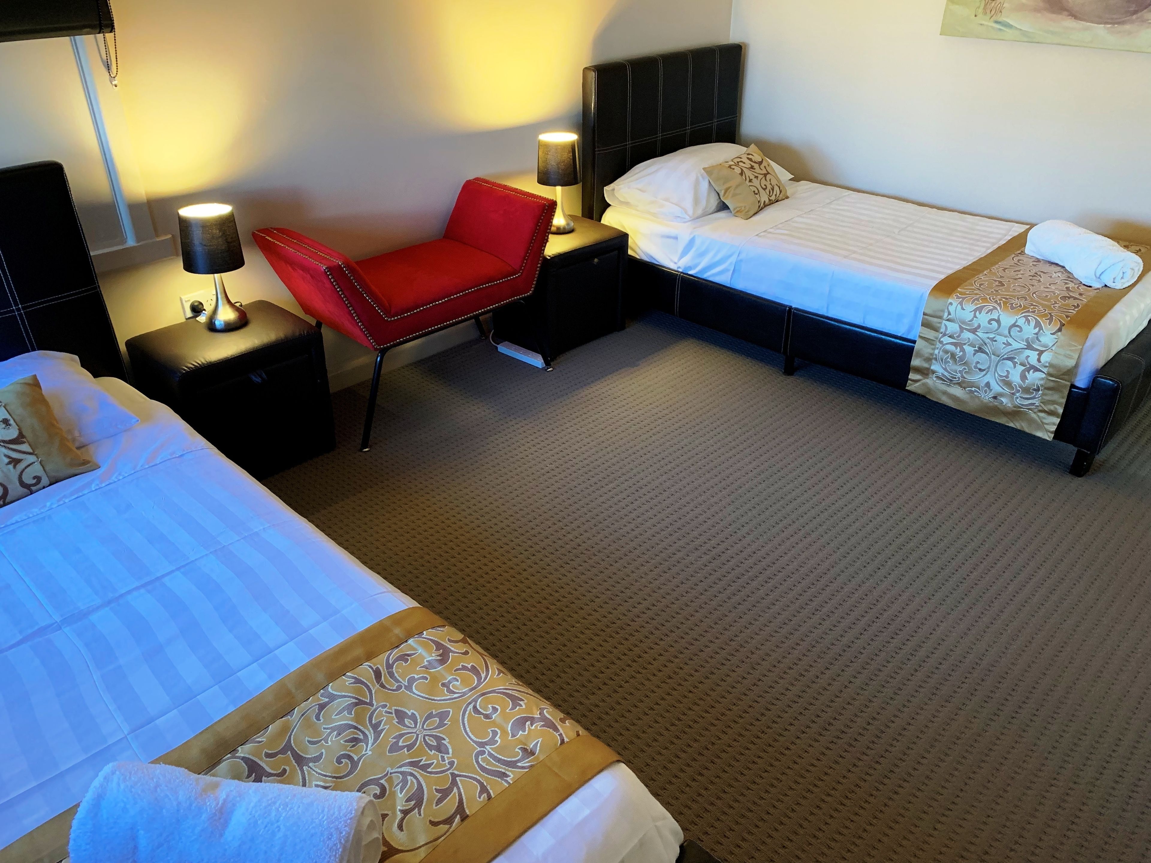 Habitación de hotel con dos camas, una silla roja, mesitas de noche con lámparas y una colcha estampada.