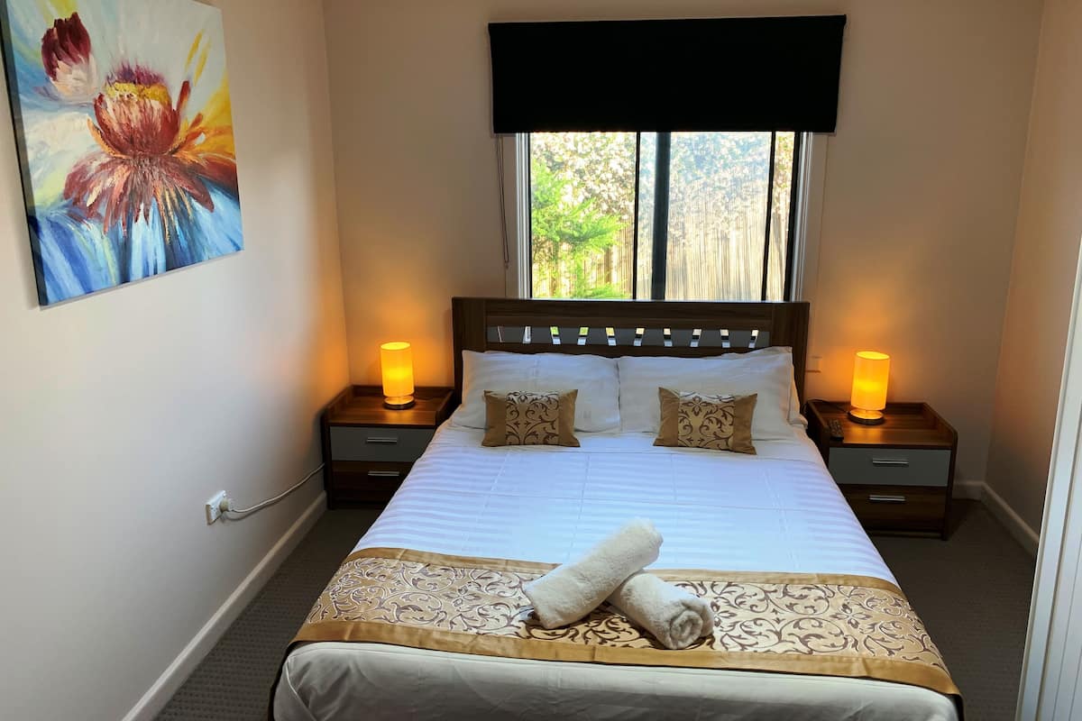 A neatly made bed with a white and gold bedspread, two bedside lamps, and a painting on the wall.