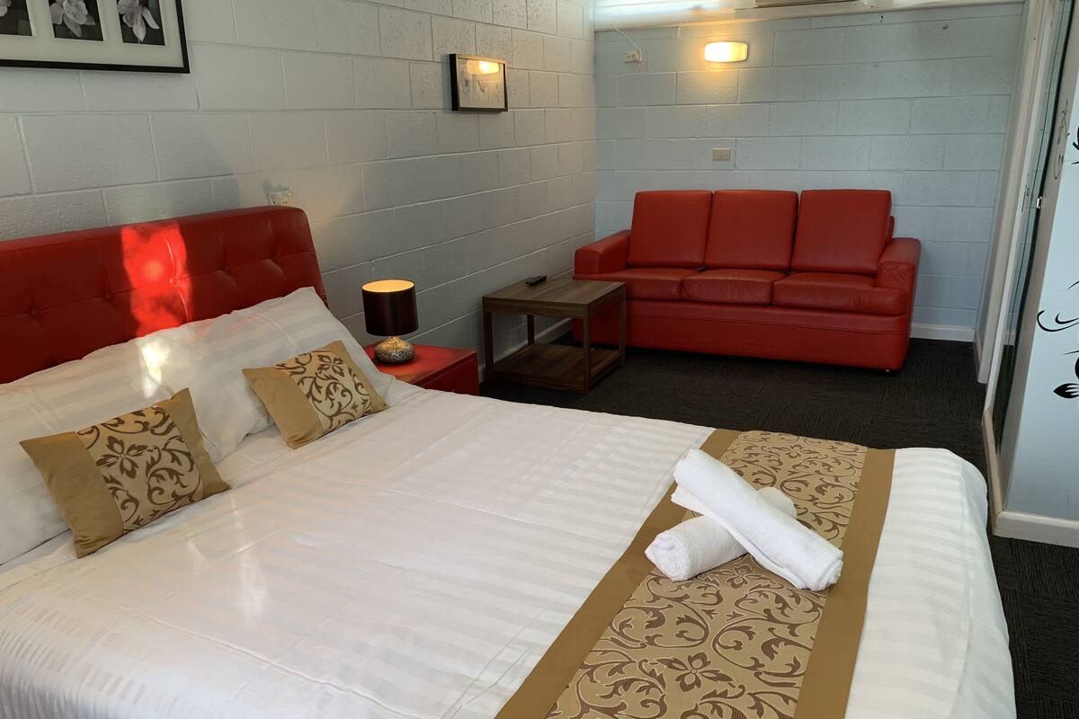 A hotel room with a large bed, red headboard, and a red sofa. There is a small table with a lamp and a picture frame on the wall.