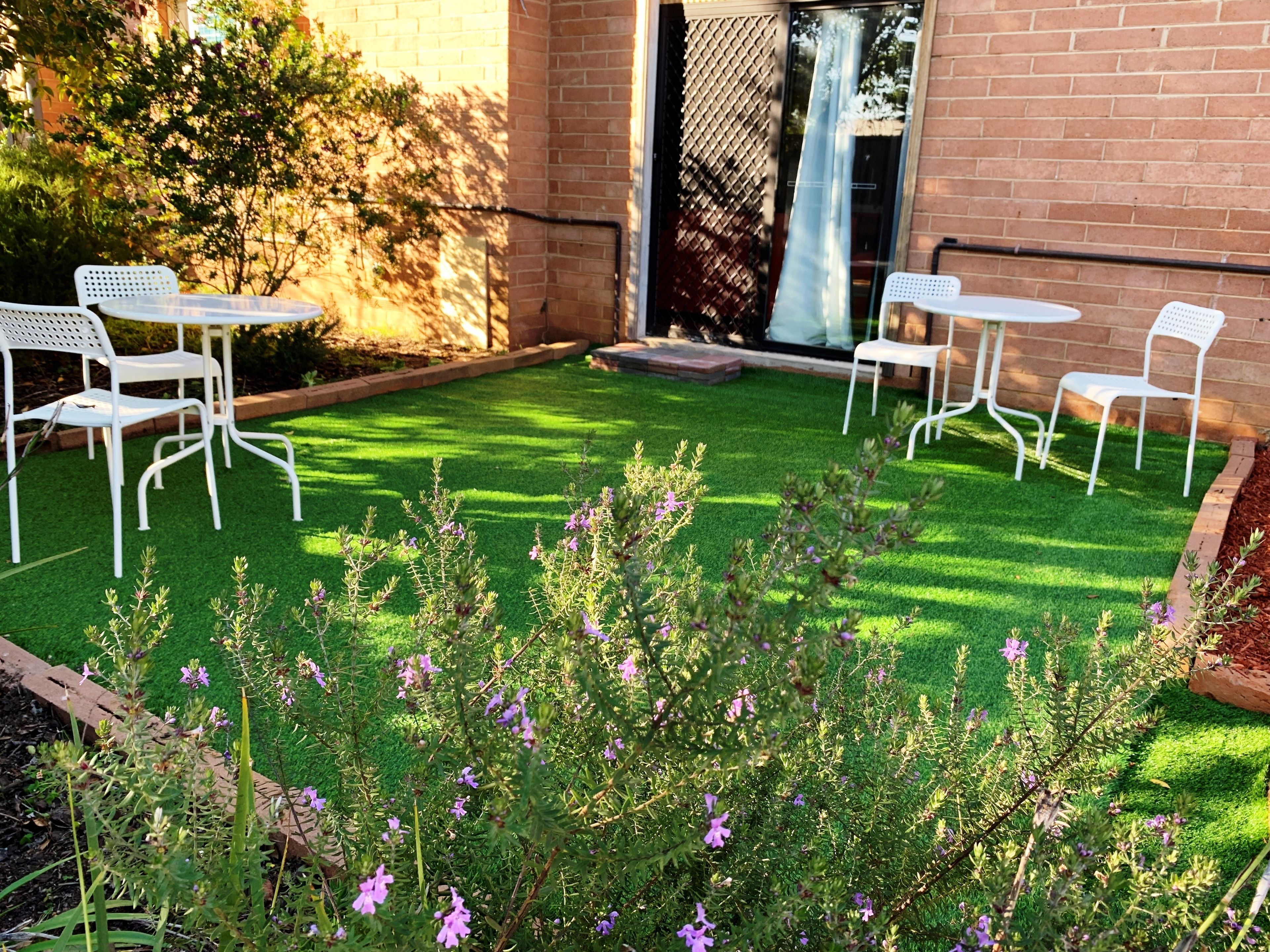 Un patio con sillas y mesas de metal blanco, rodeado de un césped cuidado y plantas con flores.