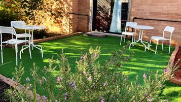 Un patio con sillas y mesas de metal blanco, rodeado de un césped cuidado y plantas con flores.