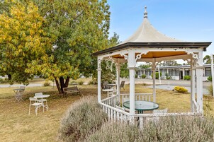A gazebo with a round table and chairs, surrounded by grass and trees.