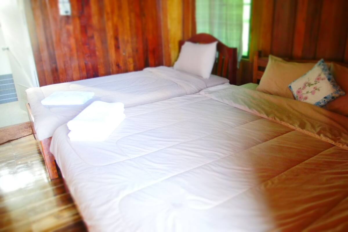 A double bed with white bedding and a floral pillow, set against a wooden wall with a window in the background.