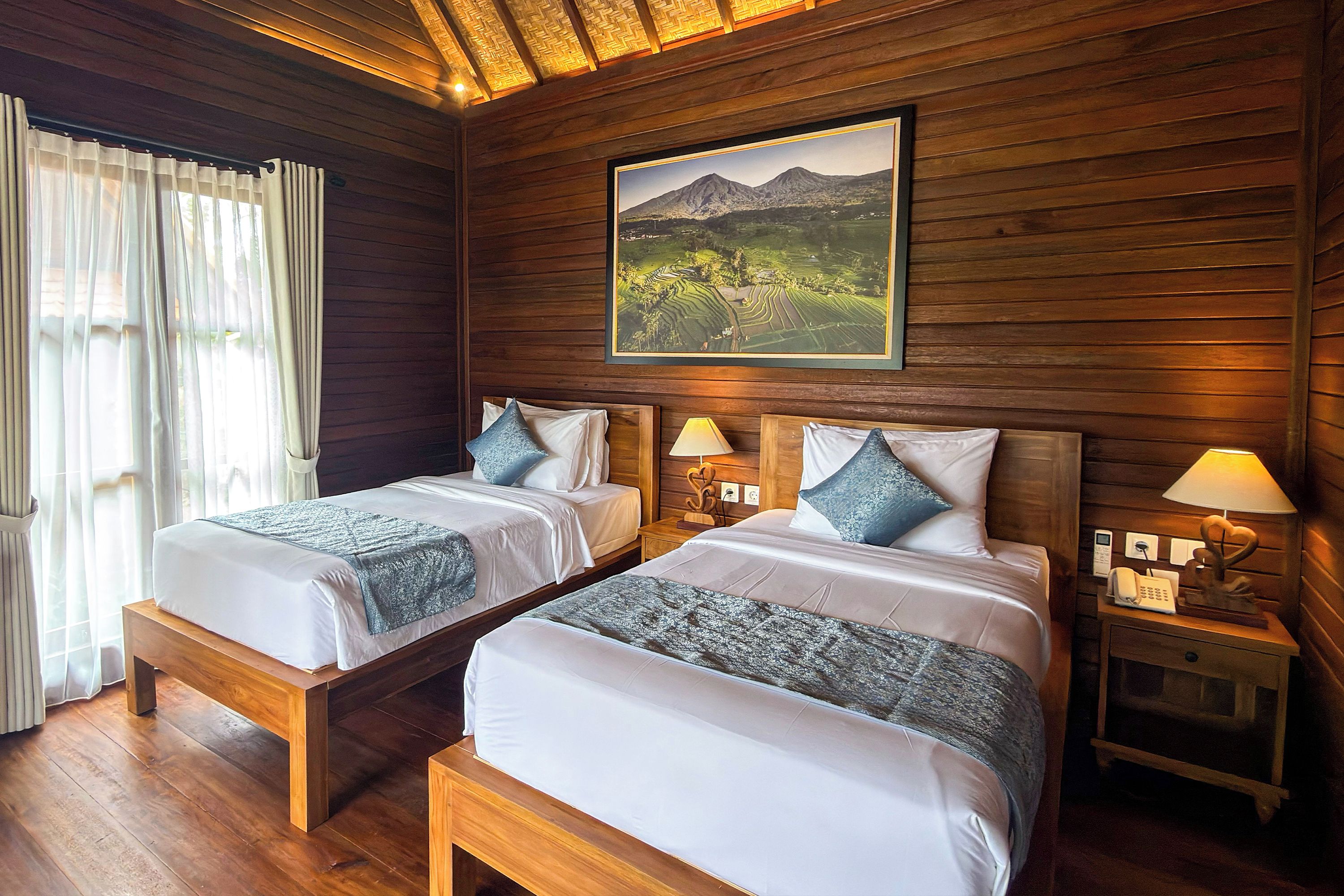 A room with two beds, wooden walls, a painting of a landscape, and bedside lamps.