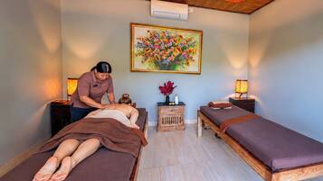 A room with two massage tables, a wooden ceiling, a wall-mounted air conditioner, and a framed floral painting.