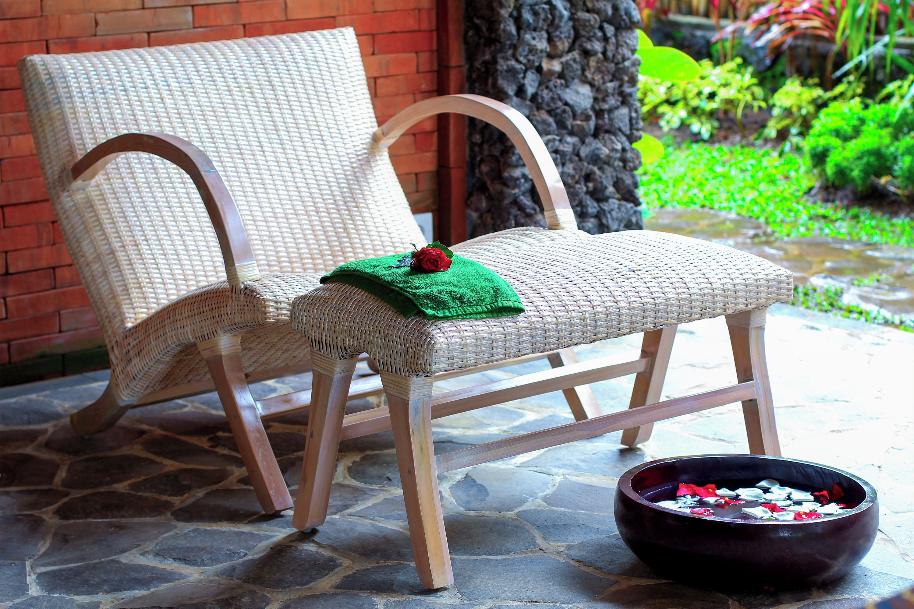 Una silla de mimbre con tela verde y una rosa roja sobre el asiento, sobre un suelo de piedra con un cuenco de pétalos de flores cerca.