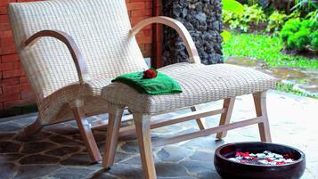 Una silla de mimbre con tela verde y una rosa roja sobre el asiento, sobre un suelo de piedra con un cuenco de pétalos de flores cerca.