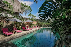 Zona junto a la piscina con sillones y sombrillas, rodeada de plantas tropicales y una piscina azul cristalina.