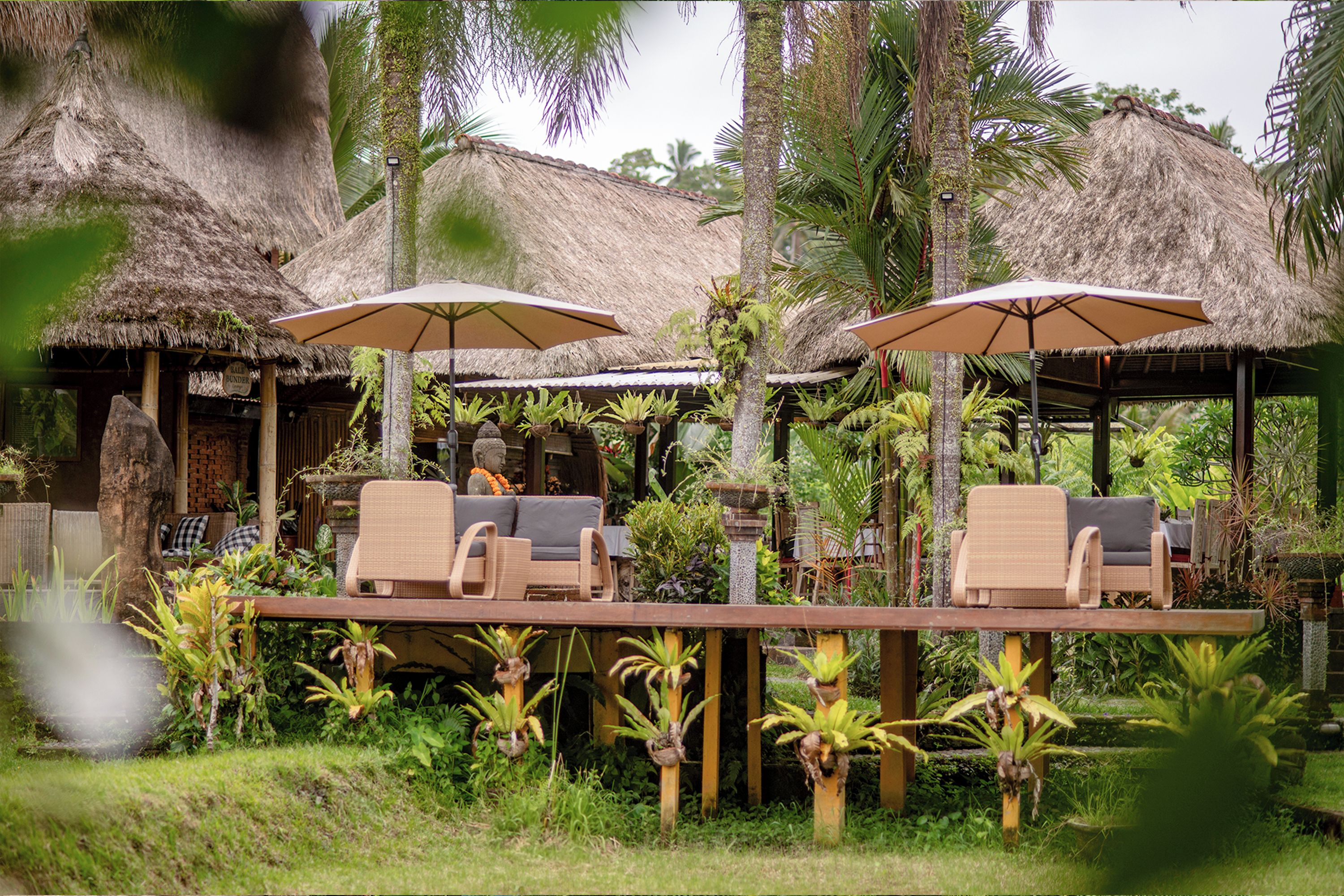 A tropical resort with thatched-roof structures, outdoor seating with umbrellas, and lush greenery.