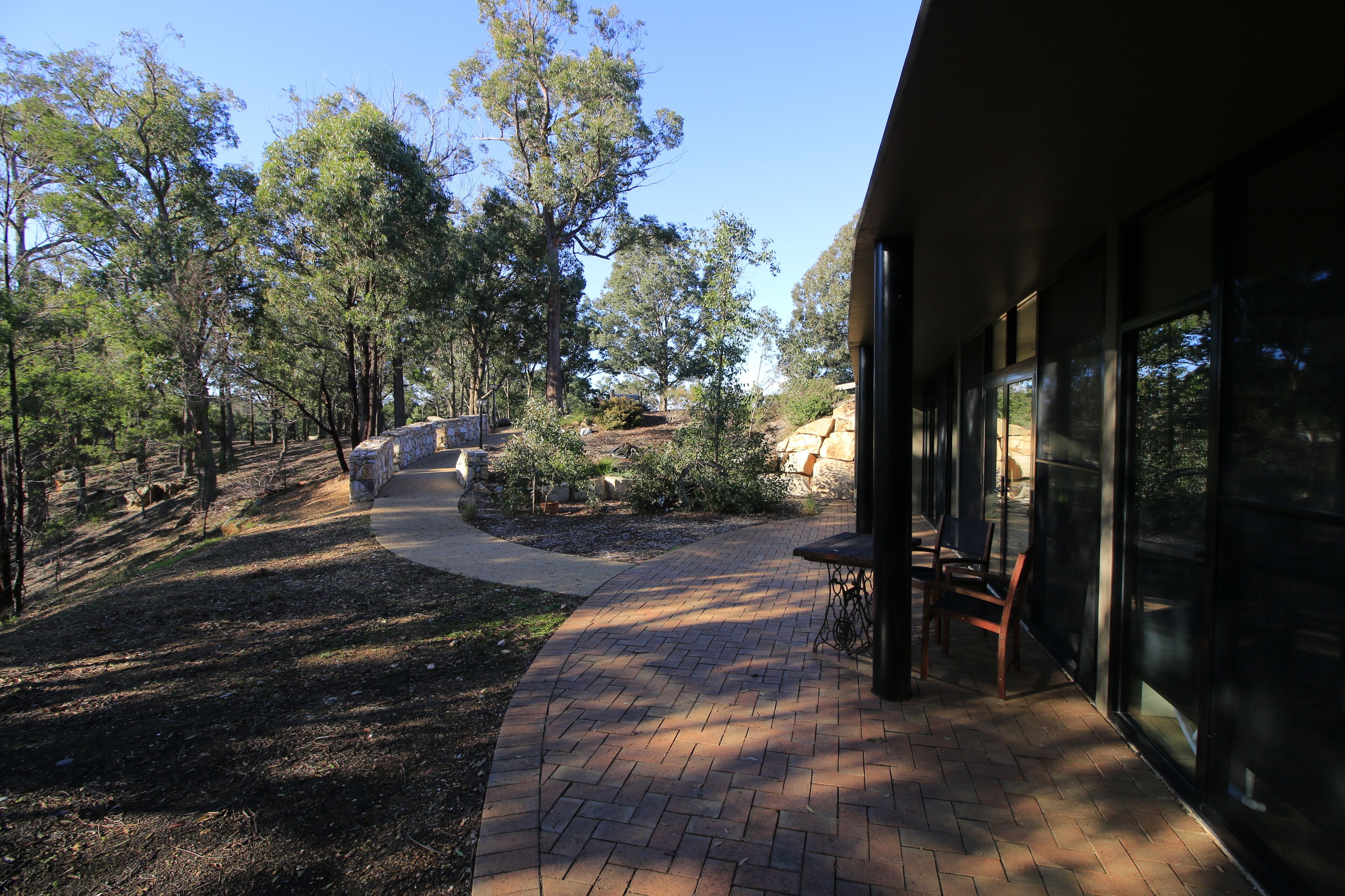 A paved pathway leading to a building with large windows, surrounded by trees and a stone wall.