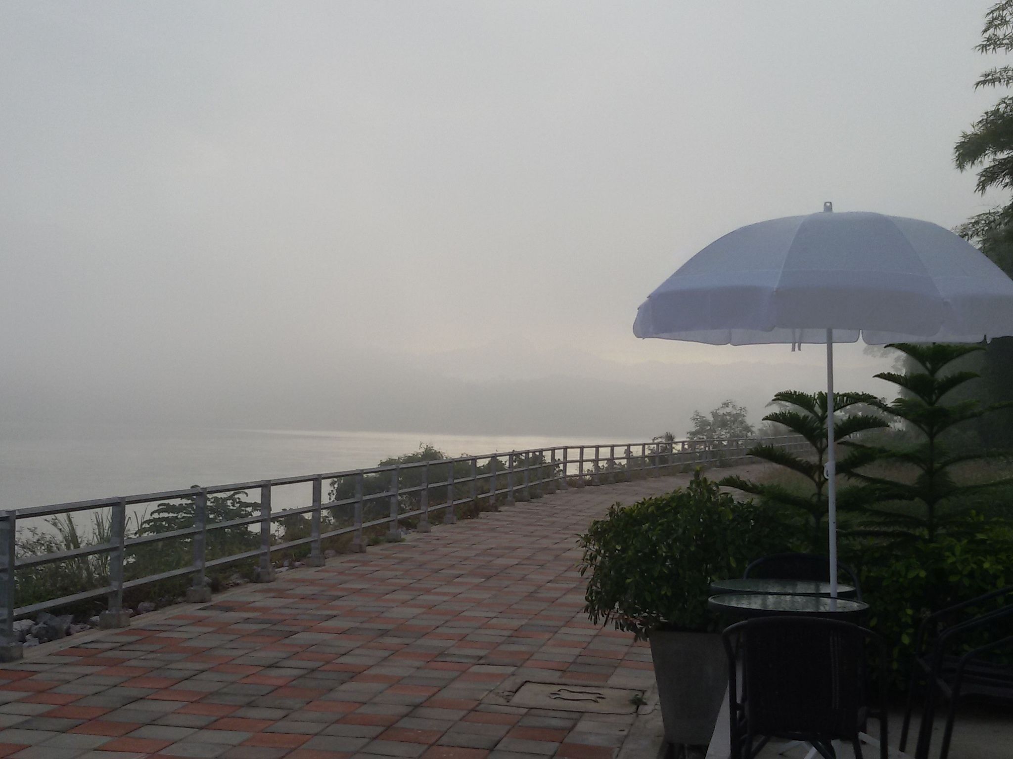 A paved walkway with a railing, a table with chairs, and a large umbrella.