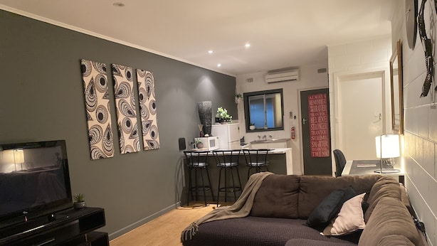 A modern living room with a TV, sofa, and coffee table. There is a kitchen area with a dining table and chairs. - Apartment on Tennyson (Port Lincoln)