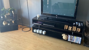 A modern living room with a flat-screen TV on a black entertainment unit, a floor lamp, and a bookshelf with various items. - Apartment on Tennyson (Port Lincoln)