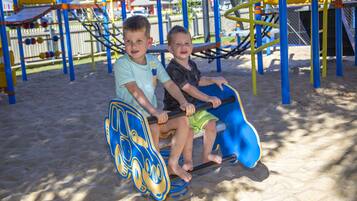 Dos niños en un juego azul y amarillo en un parque infantil con suelo de arena.