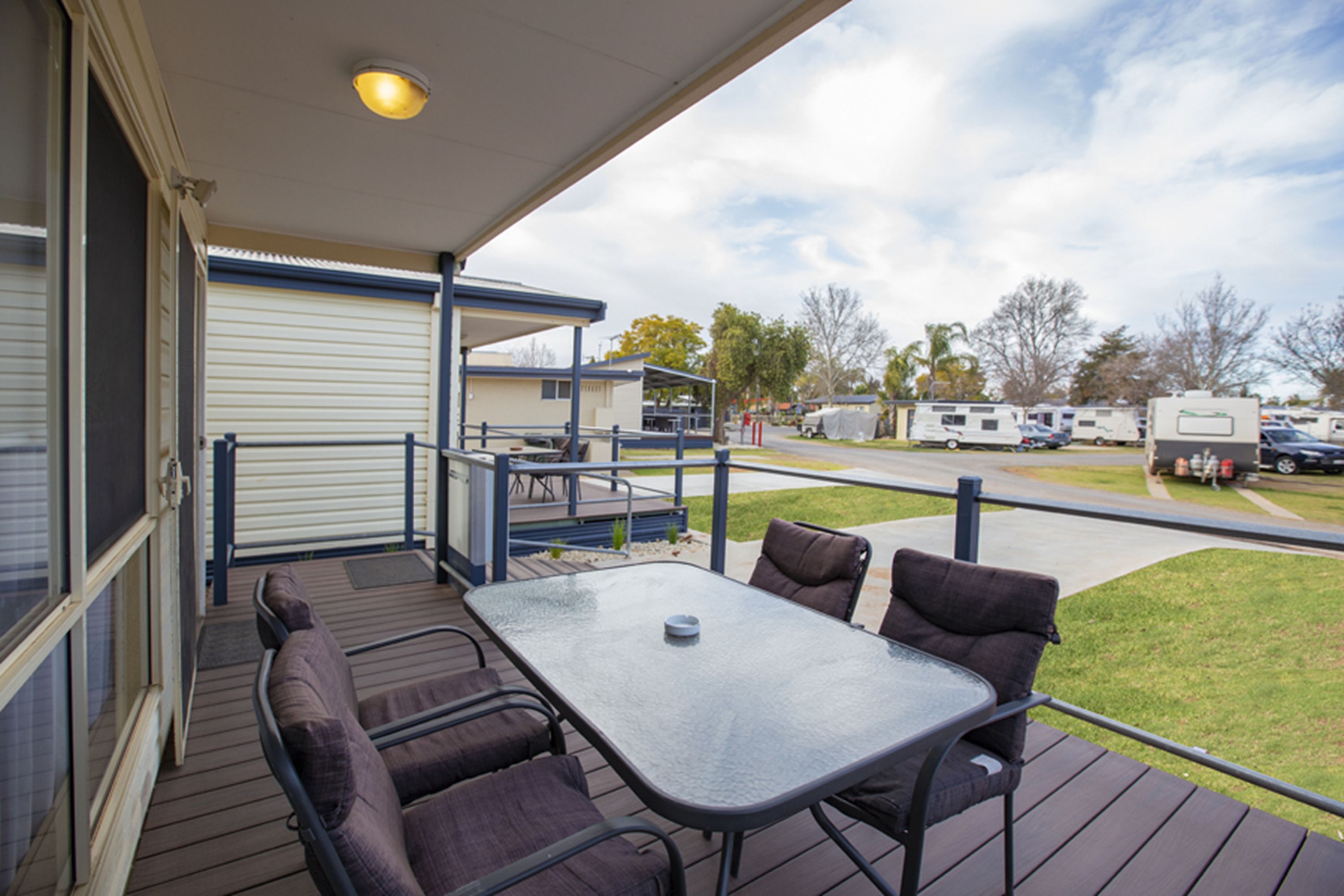 Una terraza al aire libre con una mesa y sillas, una zona cubierta con barandilla y vista a un estacionamiento con vehículos recreativos.