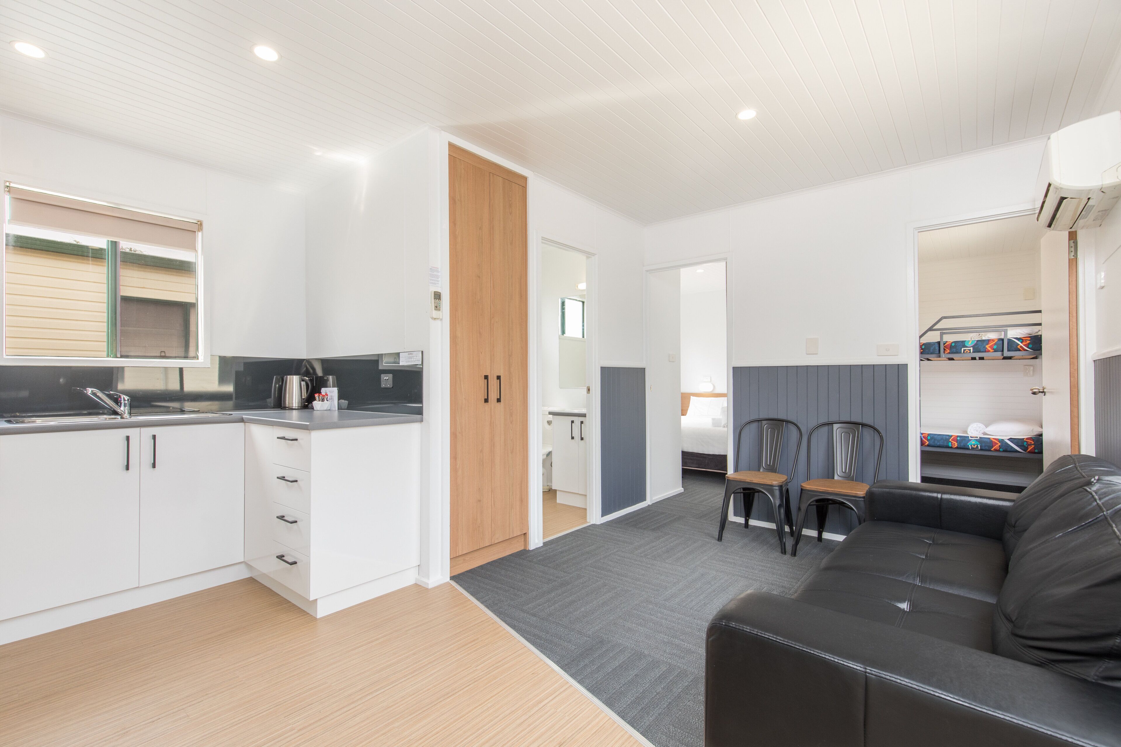 A modern kitchen with white cabinets, a black countertop, and a wooden door. There is a dining area with two chairs and a small table. A black leather sofa is visible in the foreground.