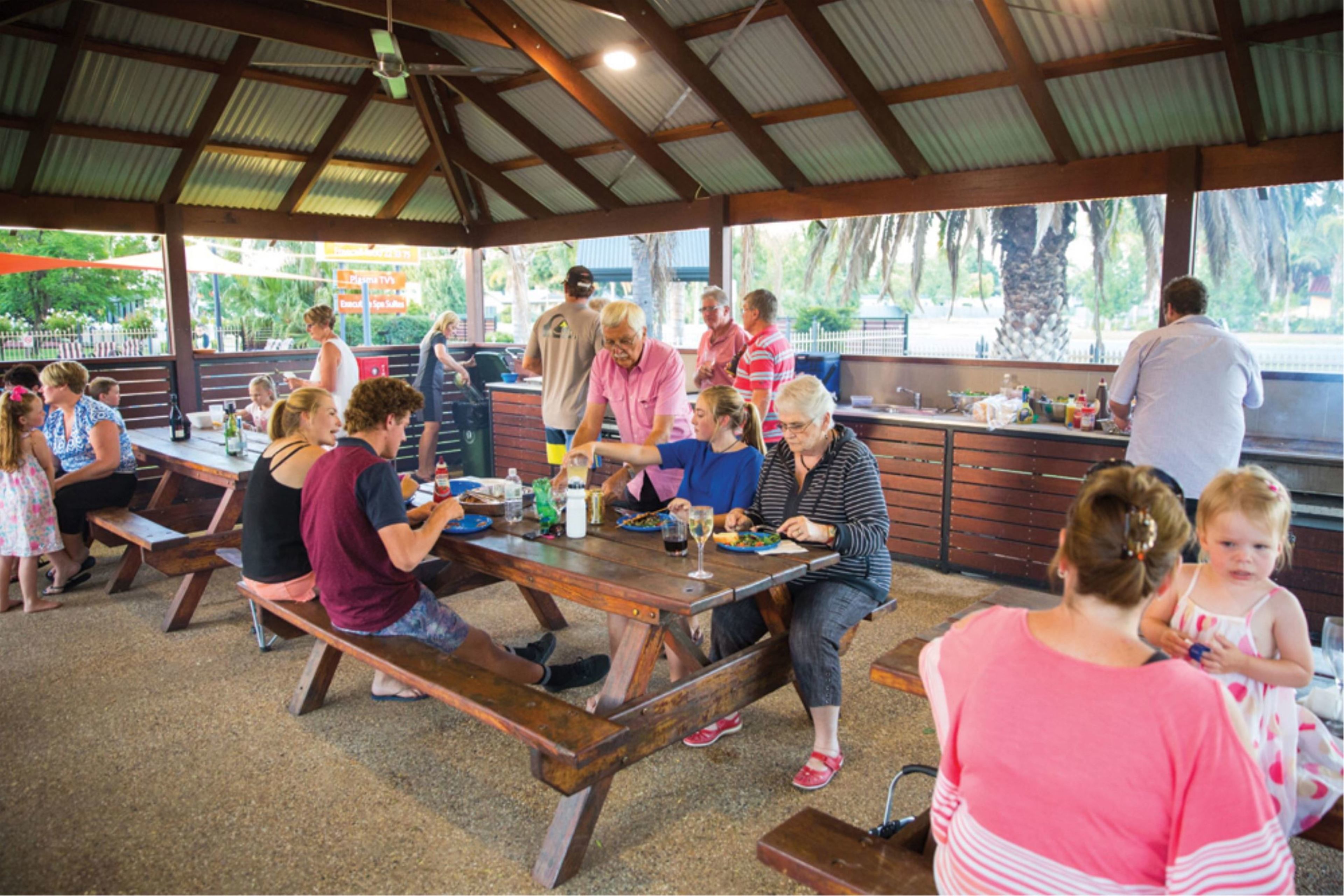 A covered outdoor dining area with wooden benches and tables, surrounded by a metal roof structure.