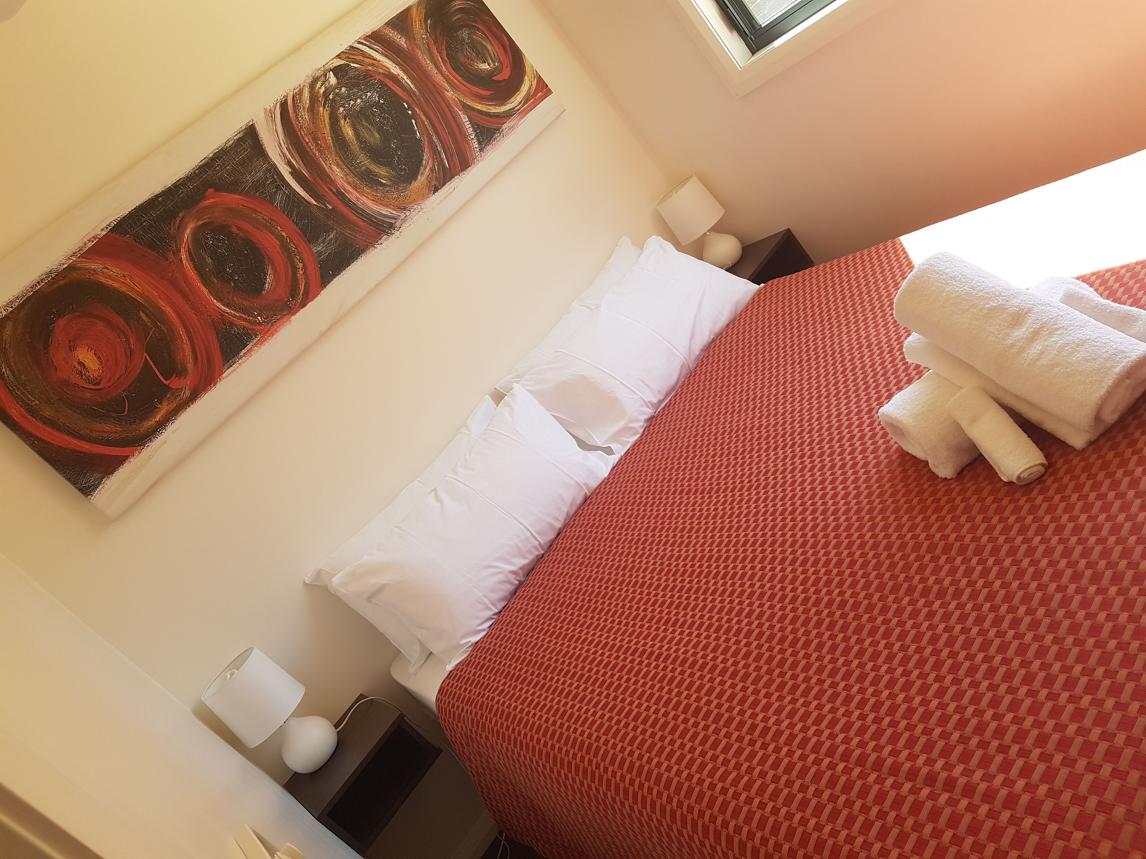 A hotel room with a bed, a painting on the wall, a lamp, and a window.