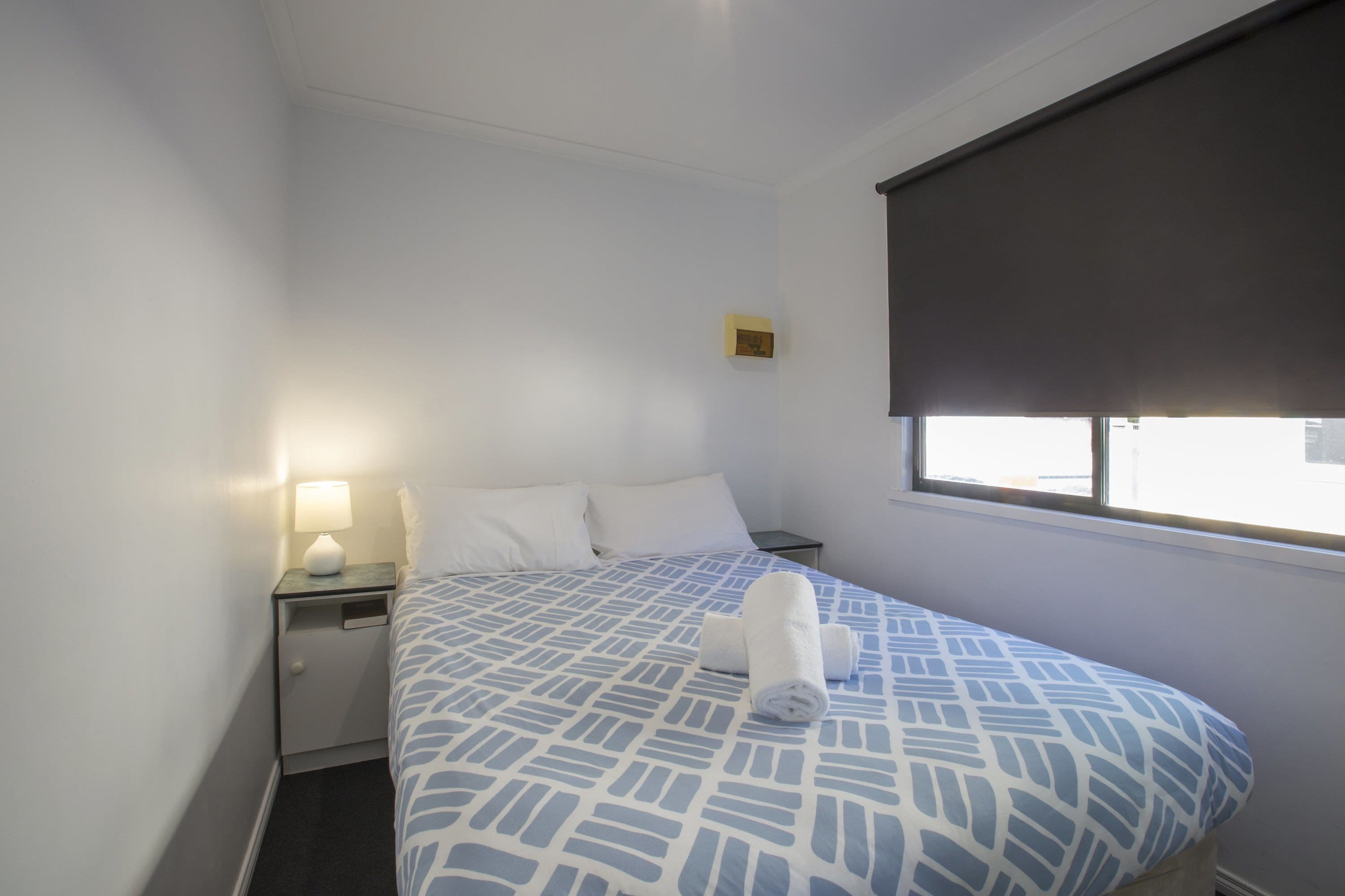 A hotel room with a bed, a nightstand, a lamp, and a window with a blind.