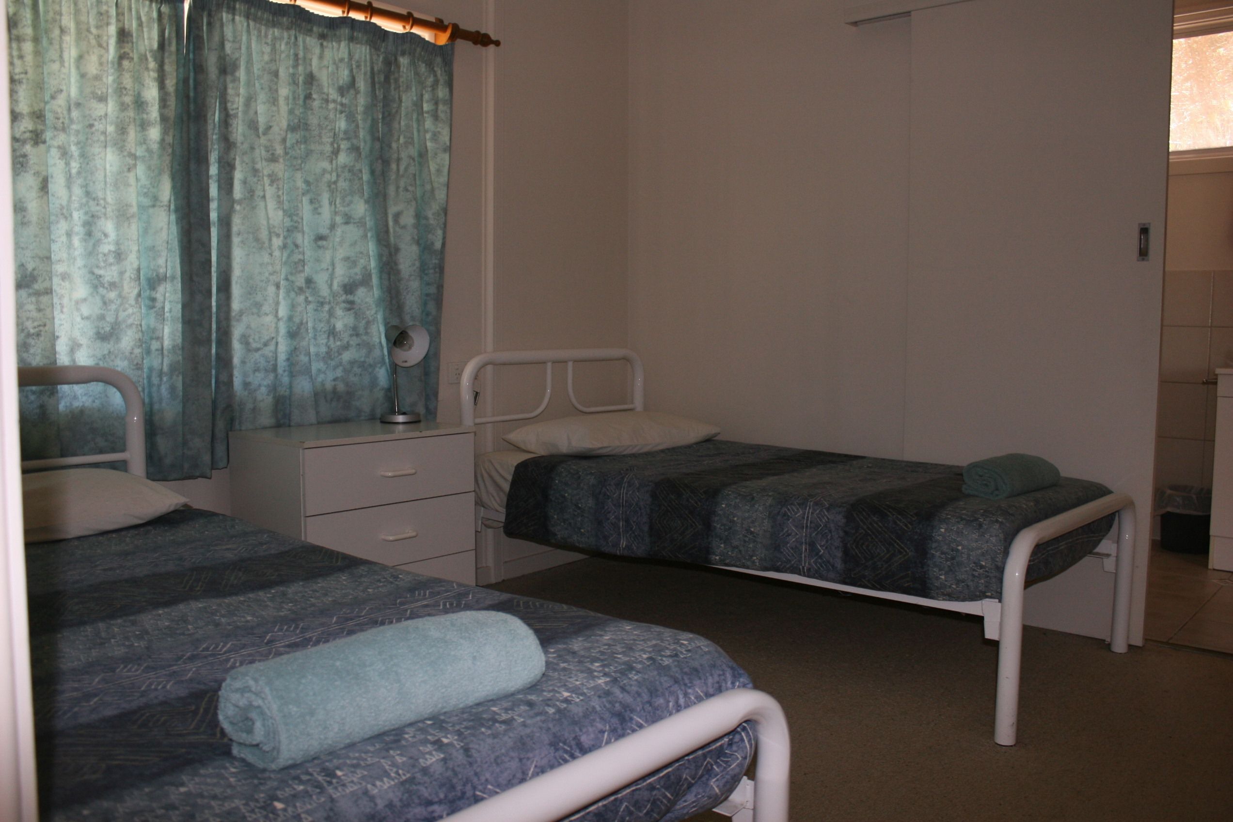 A small, simple room with two single beds, a bedside table with a lamp, and a window with curtains.