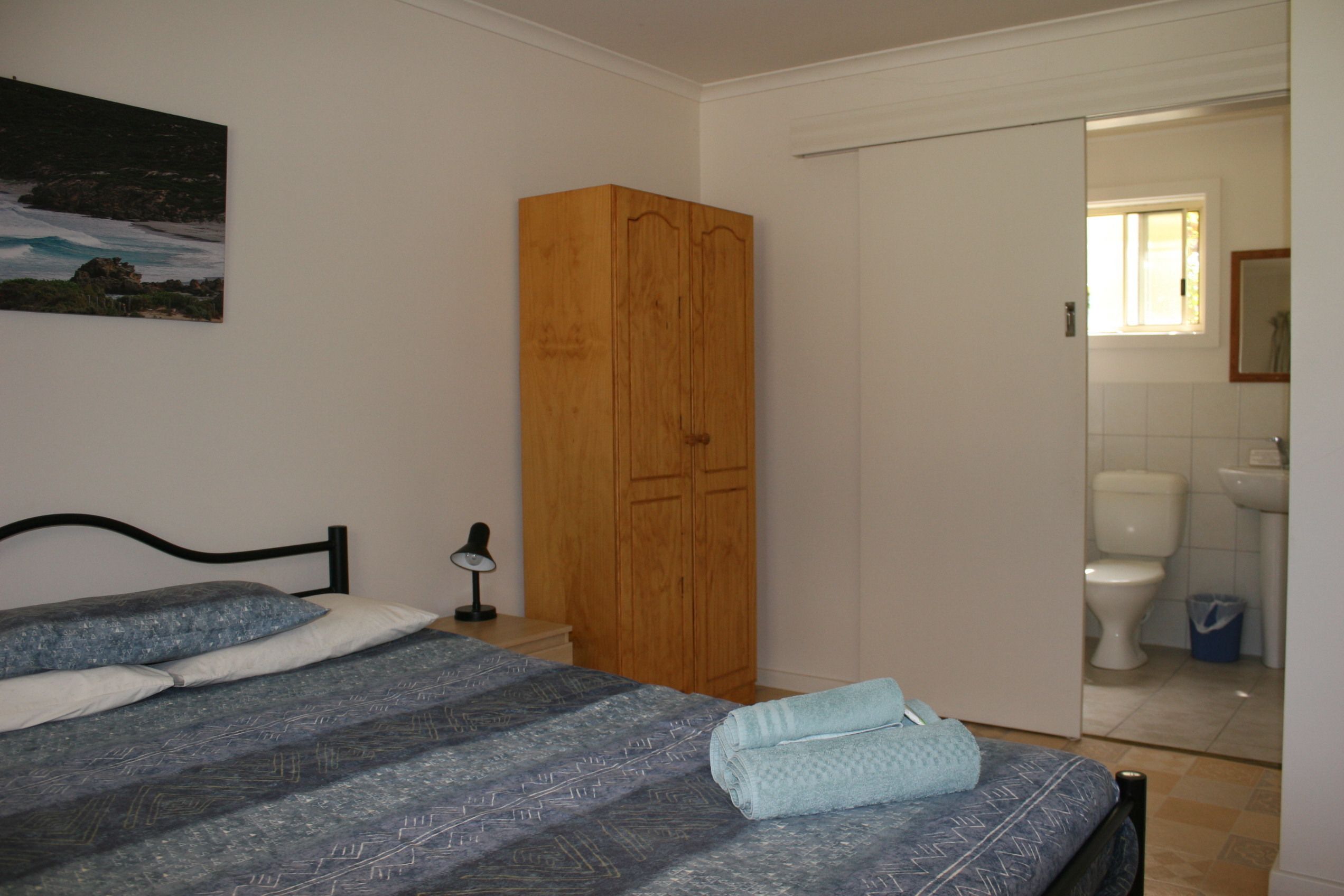 A bedroom with a bed, a wooden wardrobe, a nightstand, and a bathroom visible through an open door.