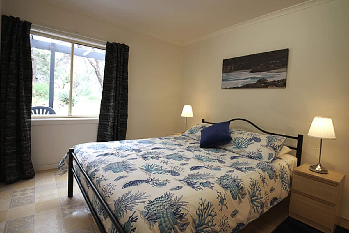 Un dormitorio con una cama, dos mesitas de noche con lámparas, una ventana con cortinas y un cuadro colgado en la pared.