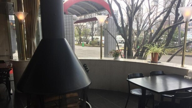 Un restaurante con una gran parrilla negra al frente, una mesa y sillas, y vista a la calle con coches y árboles.