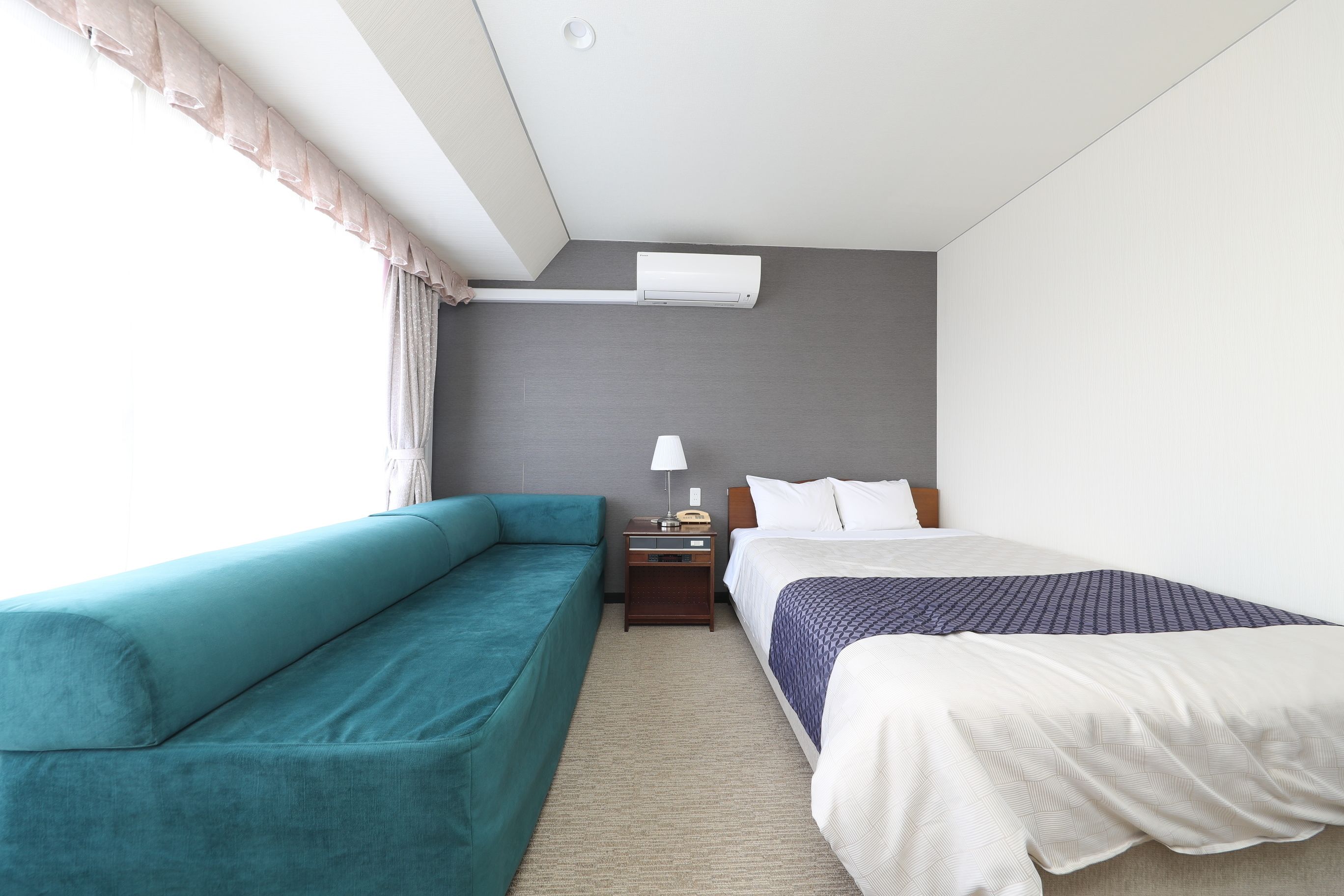 A hotel room with a teal sofa, a single bed with a patterned bedspread, a nightstand with a lamp, and an air conditioning unit on the wall.