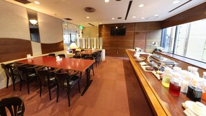 Un restaurante con una barra alargada, varias mesas y sillas. Hay una zona de bufé con diferentes opciones de comida y bebidas. La sala tiene un diseño moderno con detalles de madera y ventanas amplias.