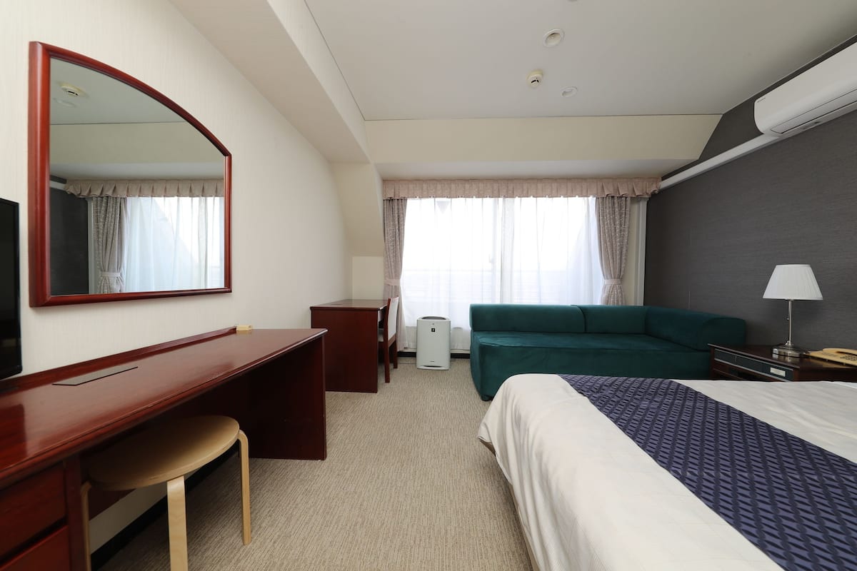 Ett hotellrum med en säng, ett skrivbord, en stol, en soffa, en tv, en lampa och en stor spegel.