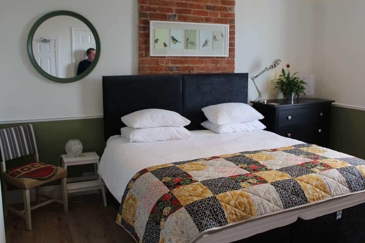 A neatly made bed with a quilt, a bedside table with a lamp, a chair, and a wall-mounted mirror.