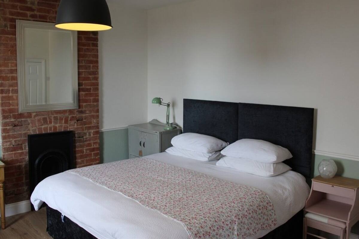 A bedroom with a bed, a nightstand, a lamp, a dresser, and a brick wall.
