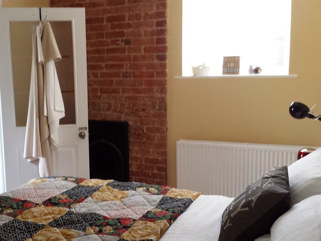 A bedroom with a quilted bedspread, a white radiator, a brick wall, and a window with a view.