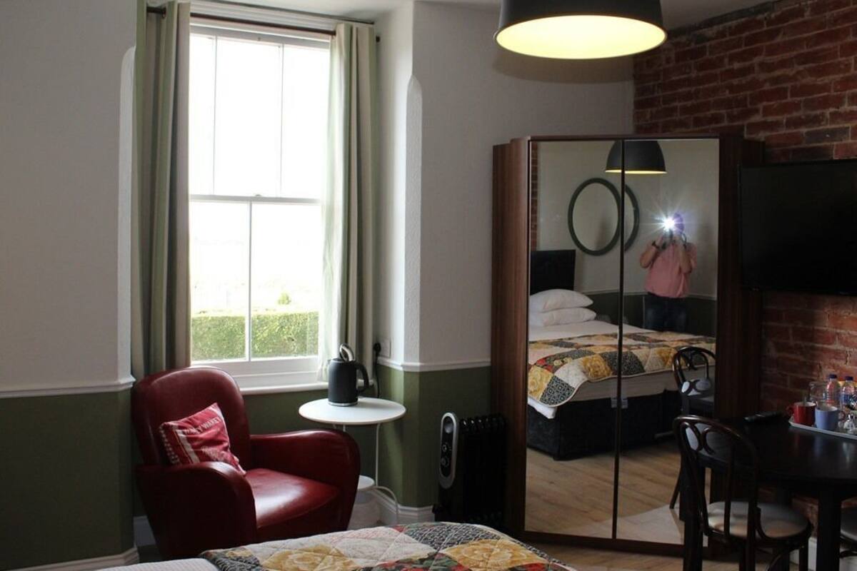 A hotel room with a bed, a red armchair, a small table, a mirror, and a brick wall.
