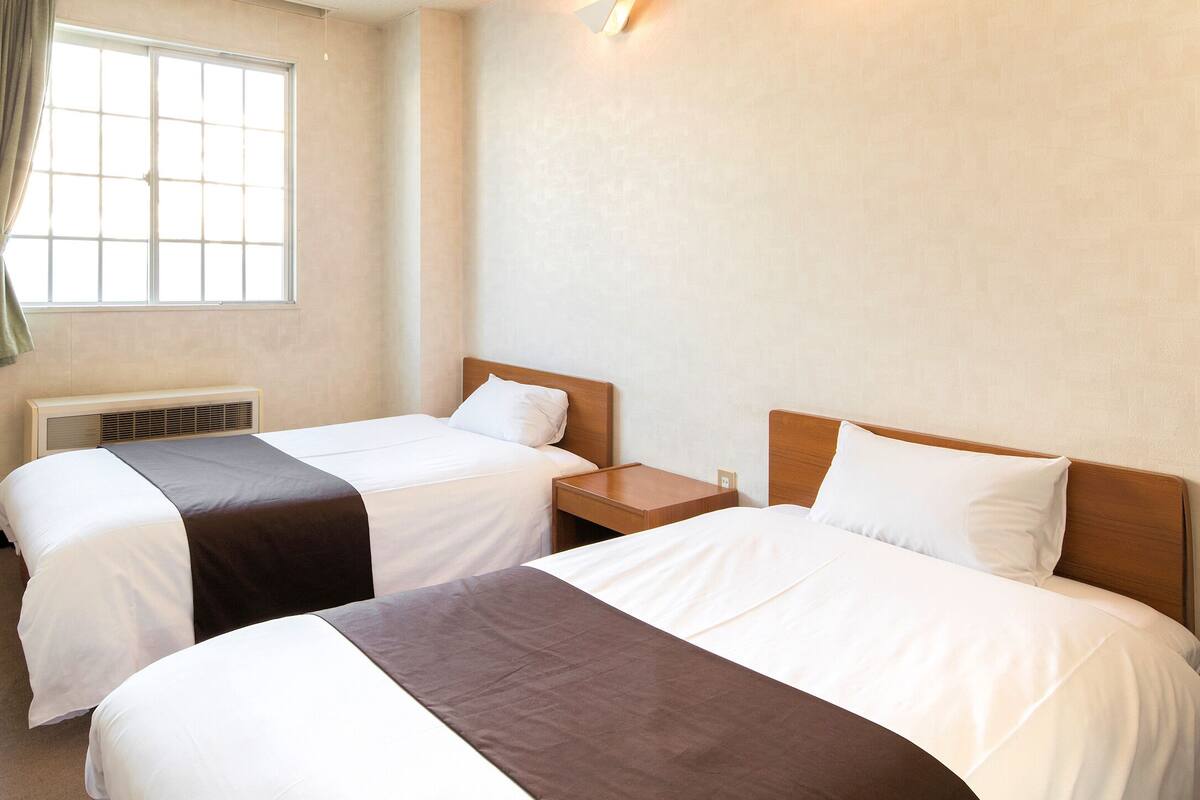 A hotel room with two beds, a window with curtains, a wooden nightstand, and a wall-mounted light fixture.