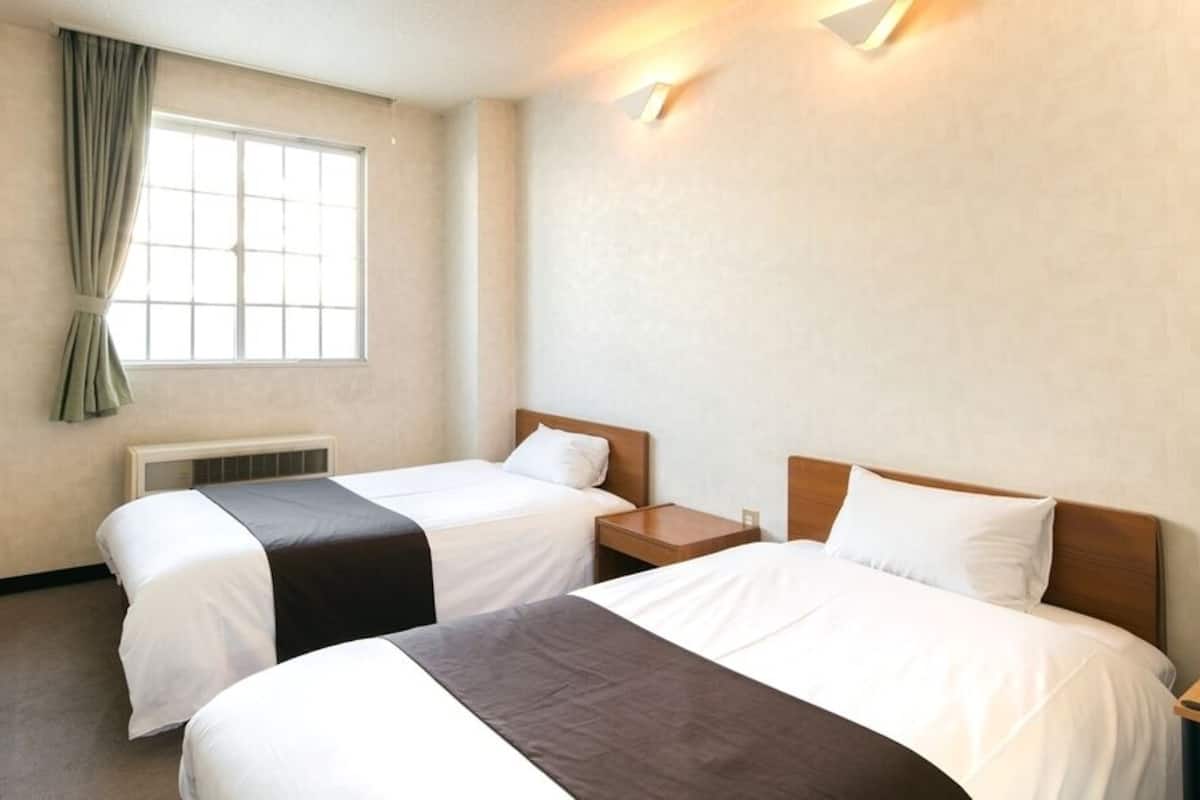 A hotel room with two beds, a window with curtains, a radiator, and a nightstand.