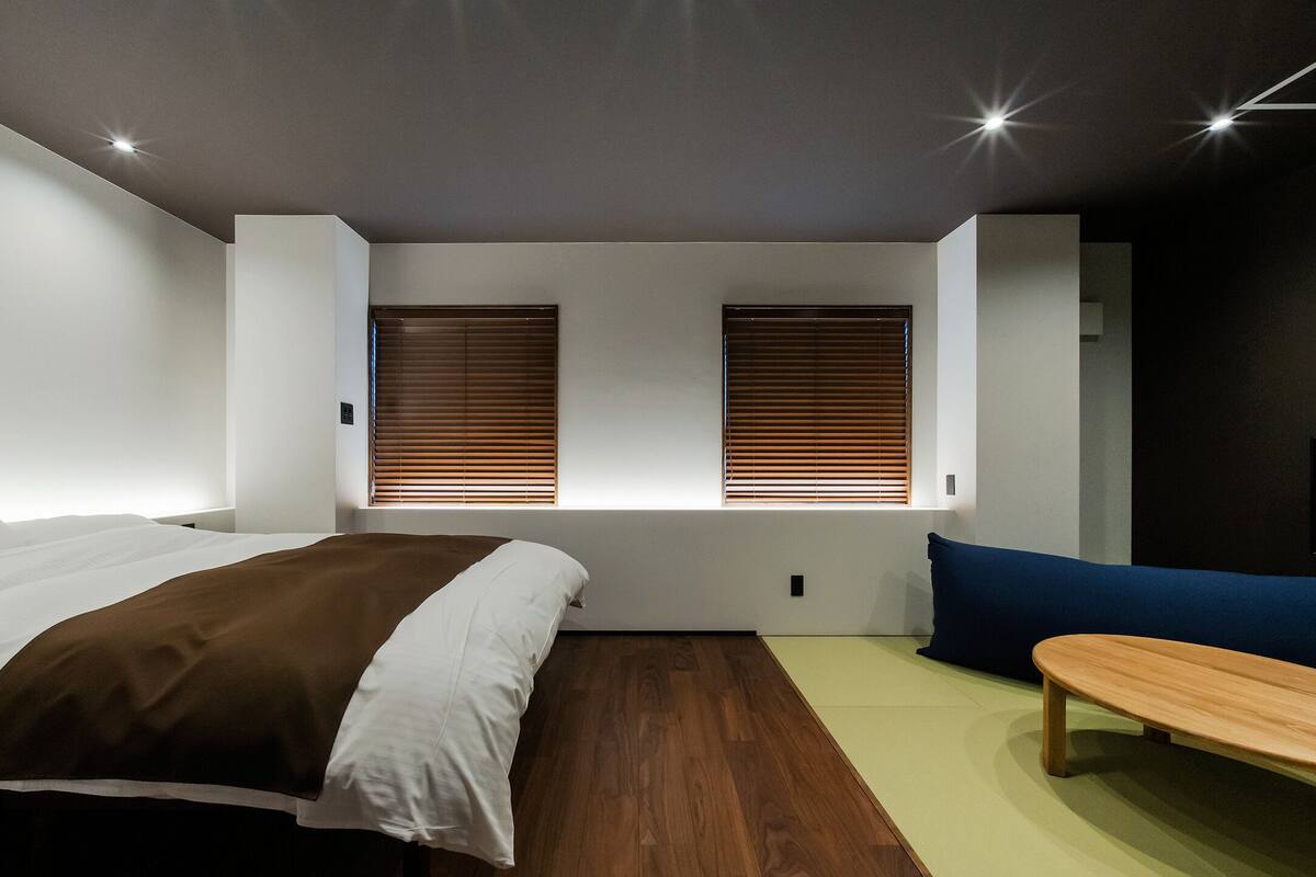 A modern bedroom with a bed, two windows with blinds, a bench, and a TV on the wall.