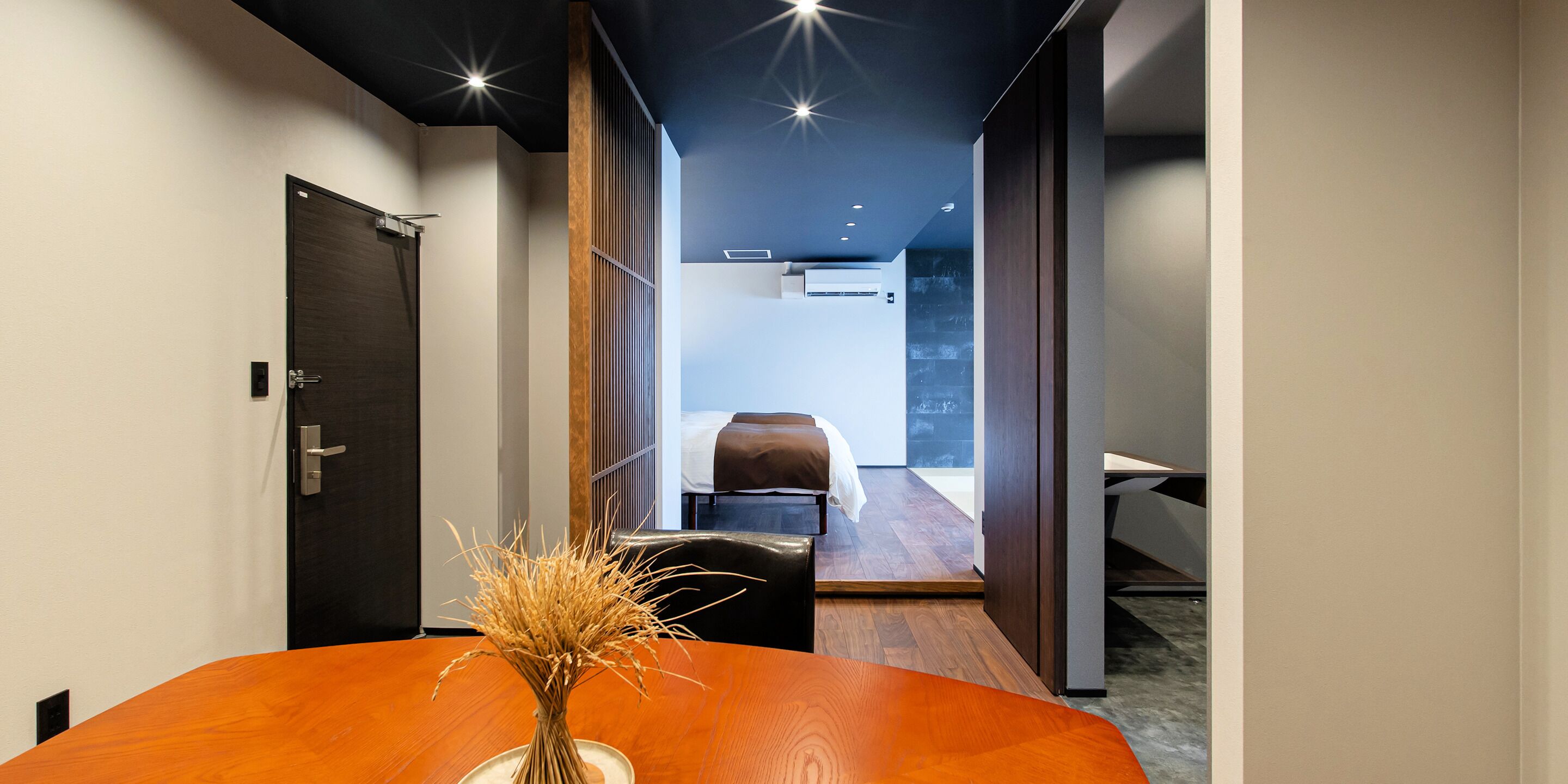 Habitación de hotel moderna con piso de madera, una cama, un escritorio y un florero con plantas secas sobre una mesa.