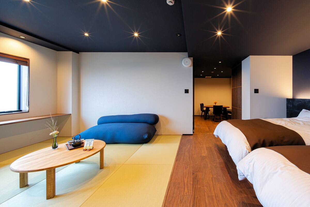 A modern hotel room with a wooden floor, a bed with white and brown bedding, a small table with a vase, and a blue sofa.