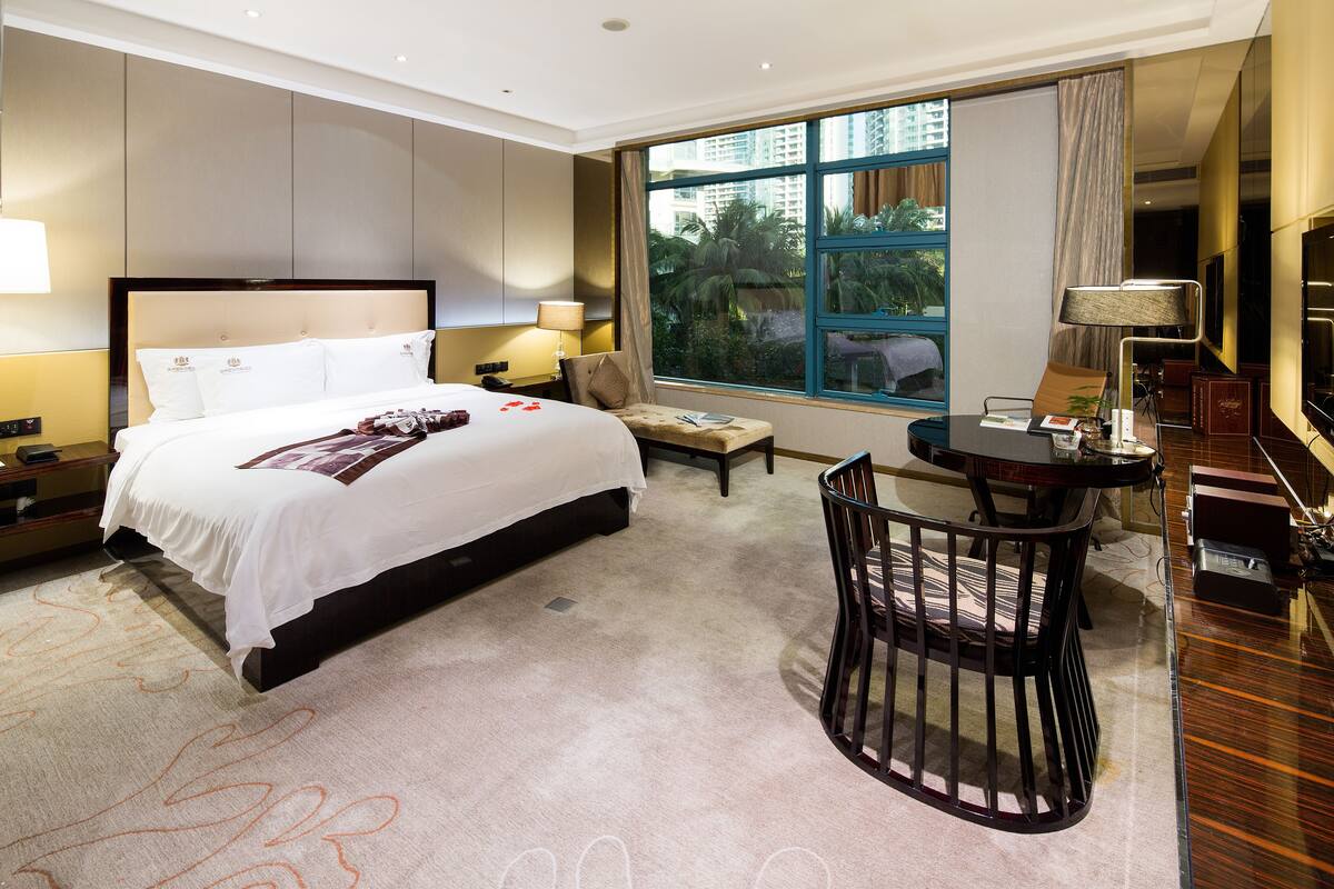 A hotel room with a large bed, a desk with a chair, a small table, and a large window offering a view of palm trees and buildings.