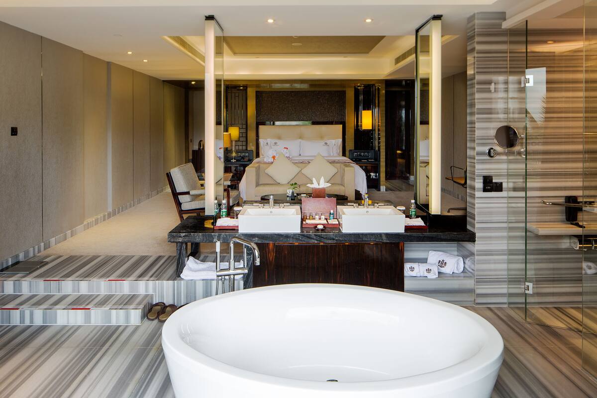A modern hotel bathroom with a large bathtub, a vanity with a mirror, and a seating area with a chair and table.