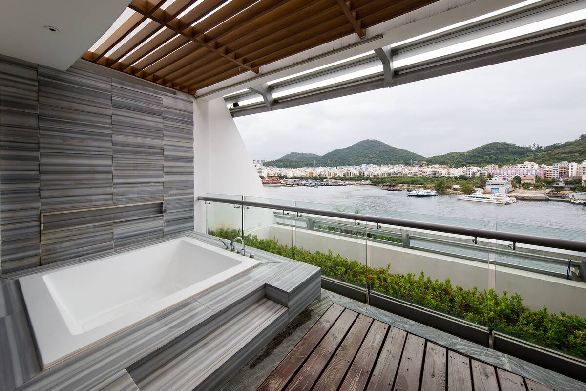 A modern balcony with a large bathtub, a wooden deck, and a view of a cityscape and mountains.