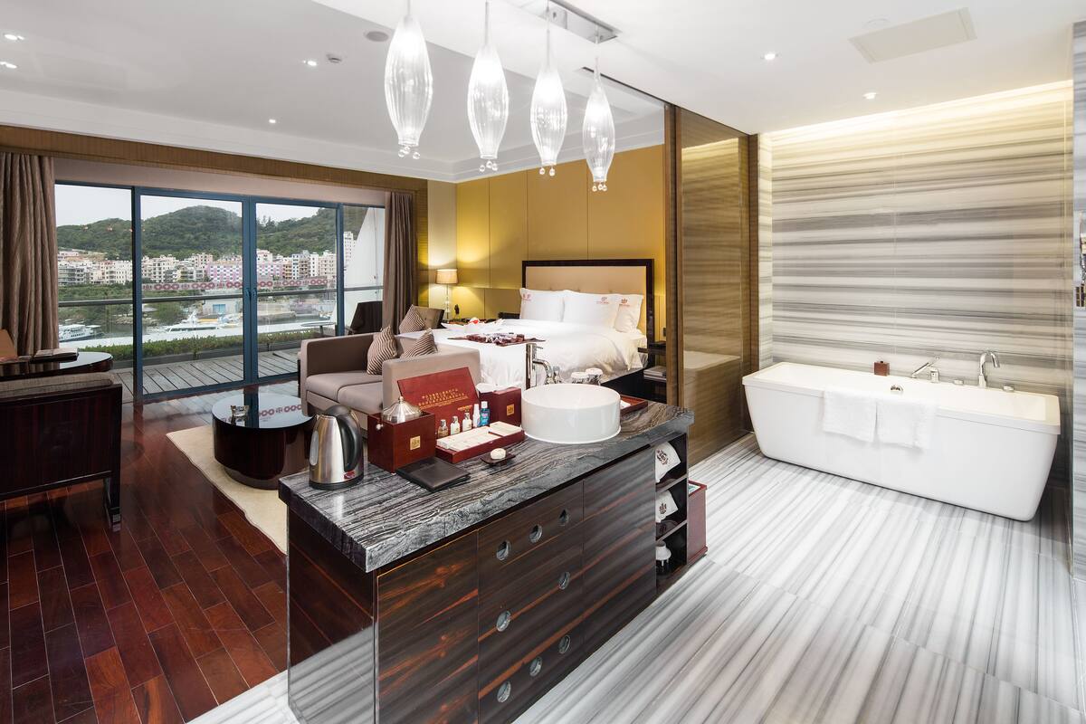 A modern hotel room with a large bed, a sitting area, a bathroom with a bathtub, and a view of the city.
