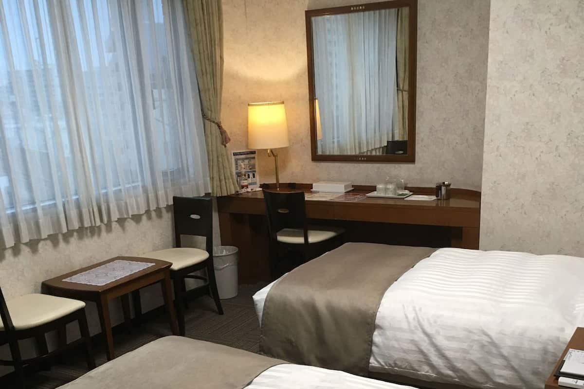 A hotel room with two beds, a desk, a chair, a lamp, and a window with curtains.