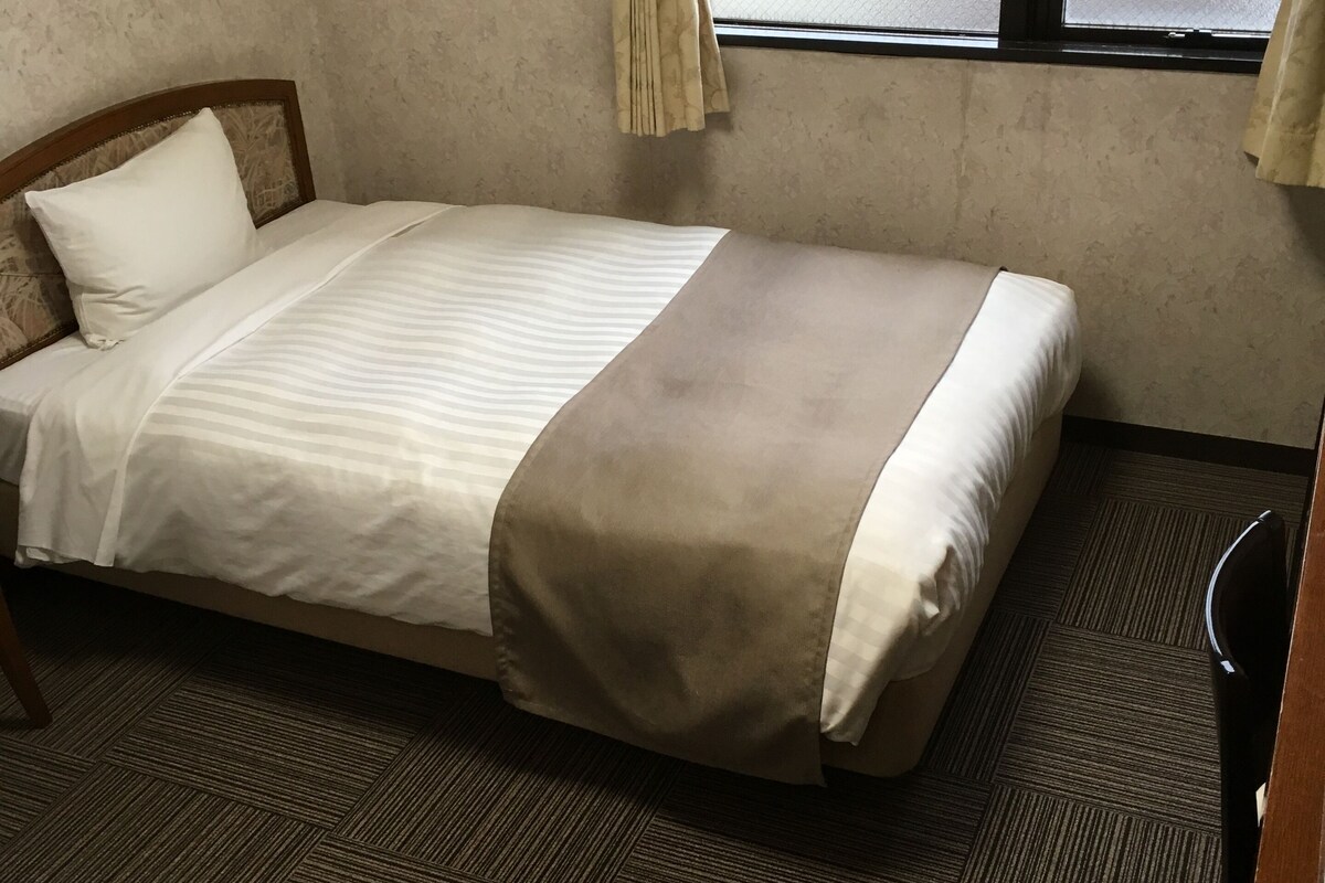 Un lit simple avec une tête de lit, une fenêtre avec des rideaux et un sol recouvert d’une moquette.