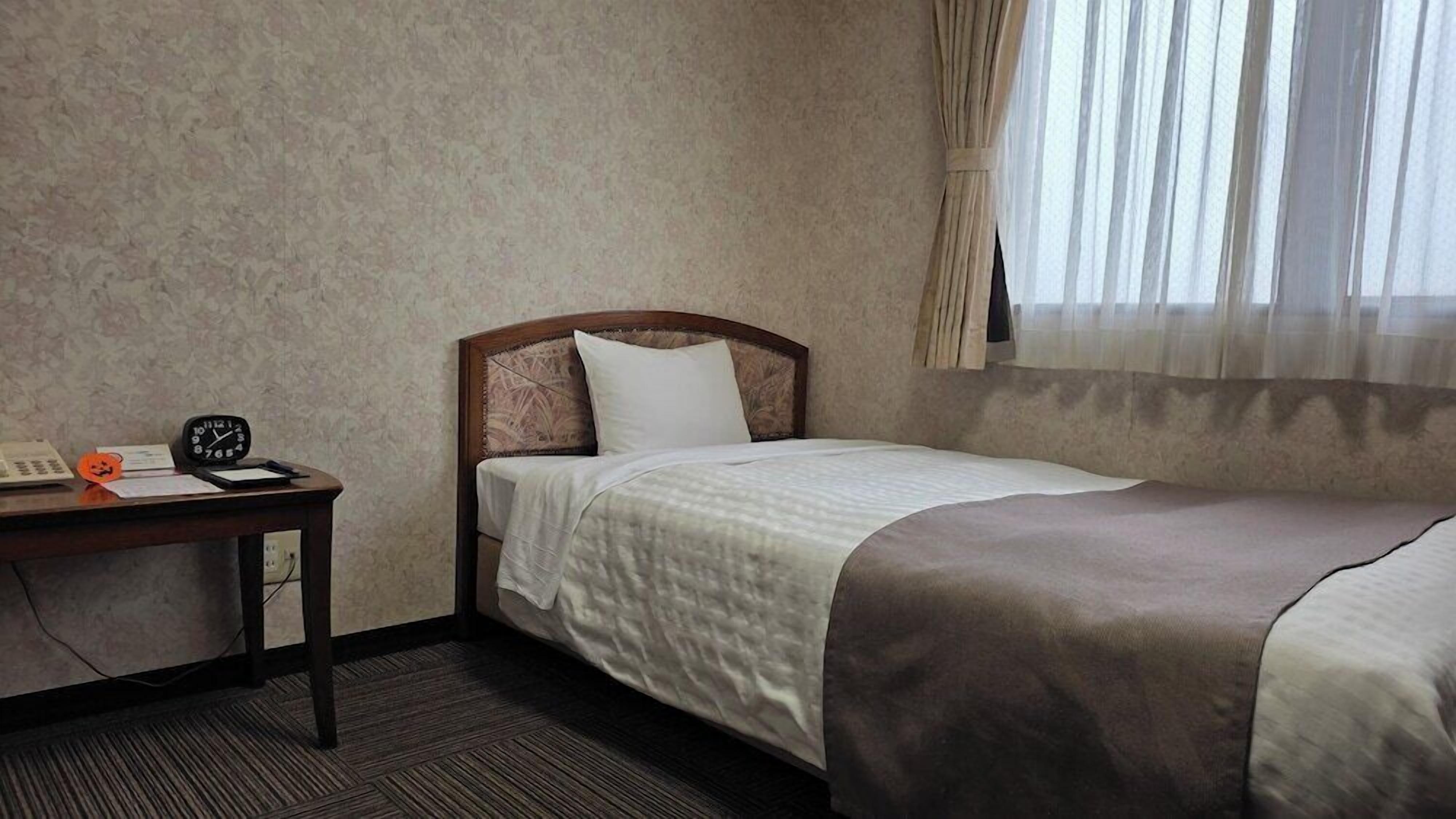 A hotel room with a single bed, a bedside table with a clock, a telephone, and a small orange decoration.