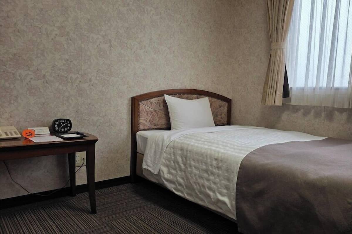 A hotel room with a single bed, a bedside table with a clock, a telephone, and a small orange decoration.