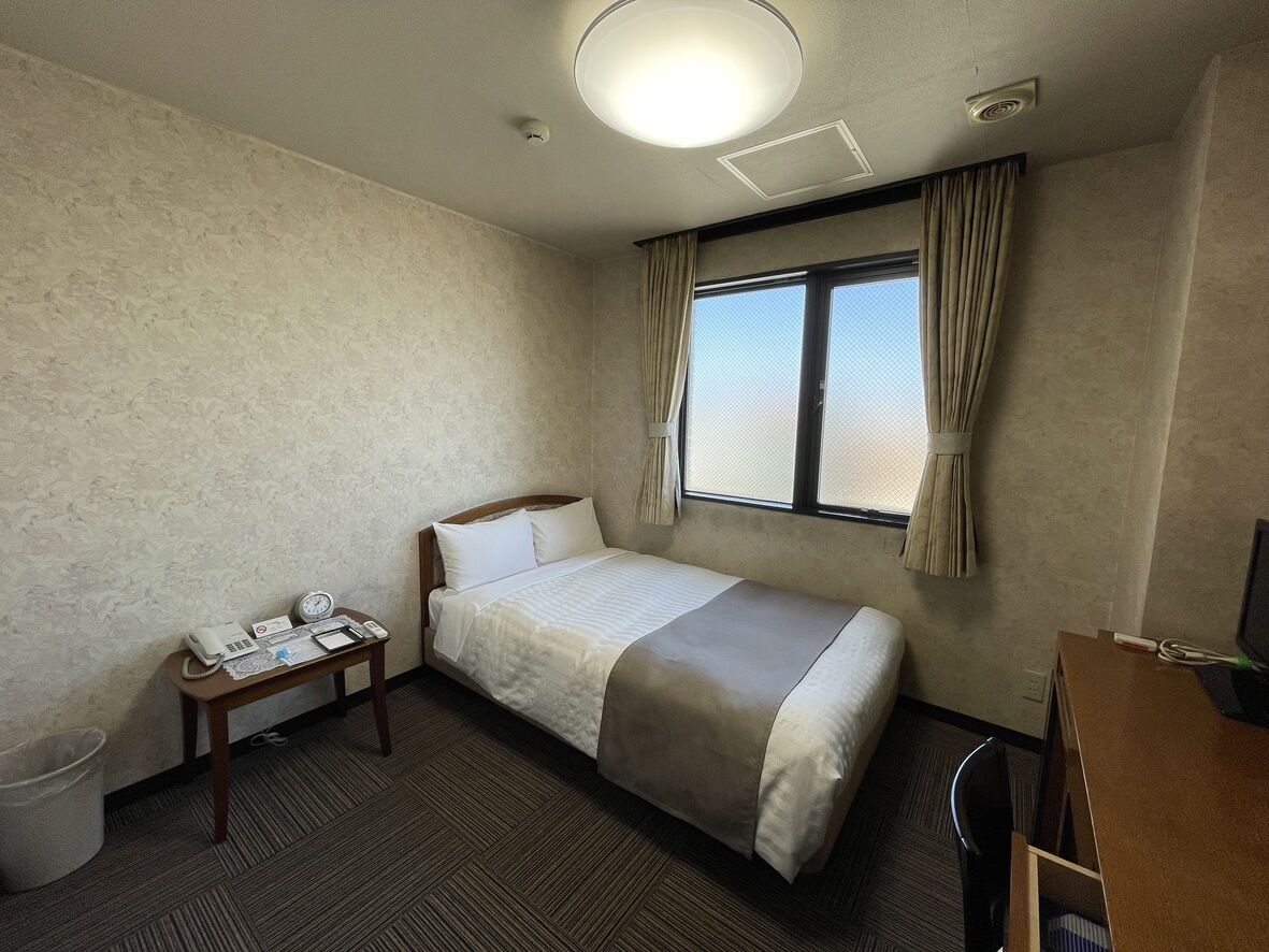 Une chambre d’hôtel avec un lit simple, une table de chevet avec un téléphone et une horloge, une fenêtre avec des rideaux et un bureau avec un ordinateur.